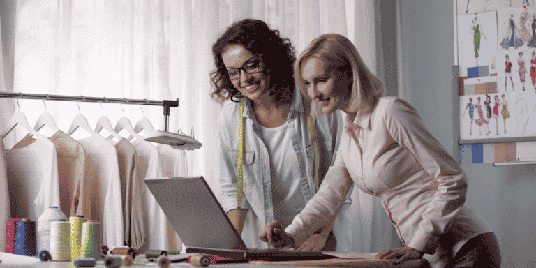 Duas mulheres, sócias, estão em pé em frente a um balcão de um ateliê de moda, usando um notebook. Ao fundo há uma arara de roupas. Como criar um site de vendas.