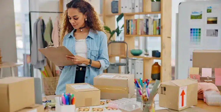 Mulher faz anotações numa prancheta; e em uma mesa há diversas caixas de papelão, em imagem que ilustra artigo sobre as ferramentas de cobrança da conta PJ da Cora.