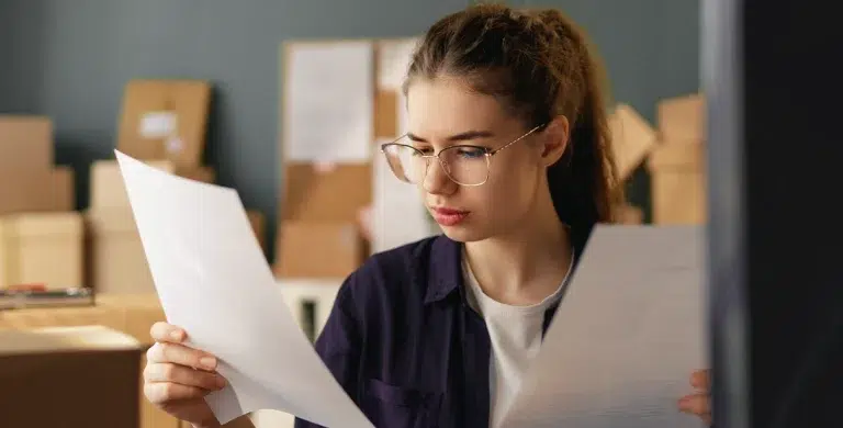 Mulher de óculos lê documentos que segura em suas mãos, em artigo sobre boletos registrados e não registrados.
