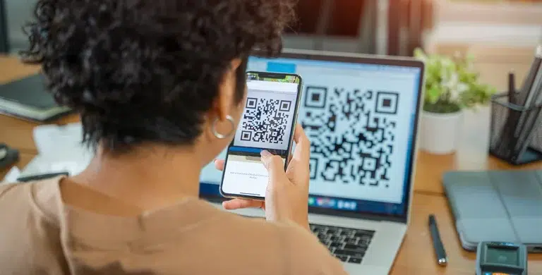 Pessoa escaneia QR code na tela de um laptop com o seu celular, em imagem que ilustra artigo sobre pagamento de boleto por Pix
