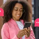 Mulher sorri ao olhar a tela do celular. Ao redor da fotomontagem há ícones de dúvida e de chat, que ajudam a ilustrar artigo sobre marketing conversacional.